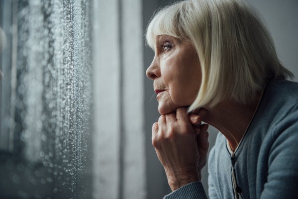 Une femme âgée aux cheveux gris fait reposer son menton sur sa main et regarde la pluie tomber par la fenêtre