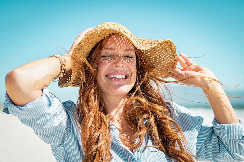 Fröhliche Frau steht lachend am Strand in der Sonne und hält ihren Sonnenhut fest.