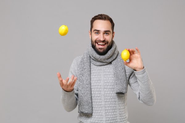 Junger Mann im grauen Pullover mit Schal posiert vor grauem Wandhintergrund und h&auml;lt Zitronen als Vitamin-C-Quelle in den H&auml;nden.