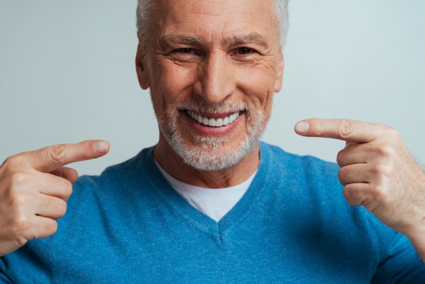 Foto von einem Senior mit blauem Pullover, der mit den Fingern auf seinen lachenden Mund zeigt.