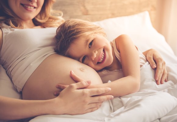 Schwangere Mutter kuschelt mit gr&ouml;sserem Kind im Bett